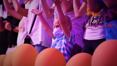 Bawiąca Się Publiczność. Mały chłopczyk w maseczce klaszcze. Przed dzieckiem ustawione w rzędzie pomarańczowe balony