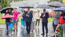 Uczestnicy wydarzenia stoją pod parasolami, pada deszcz